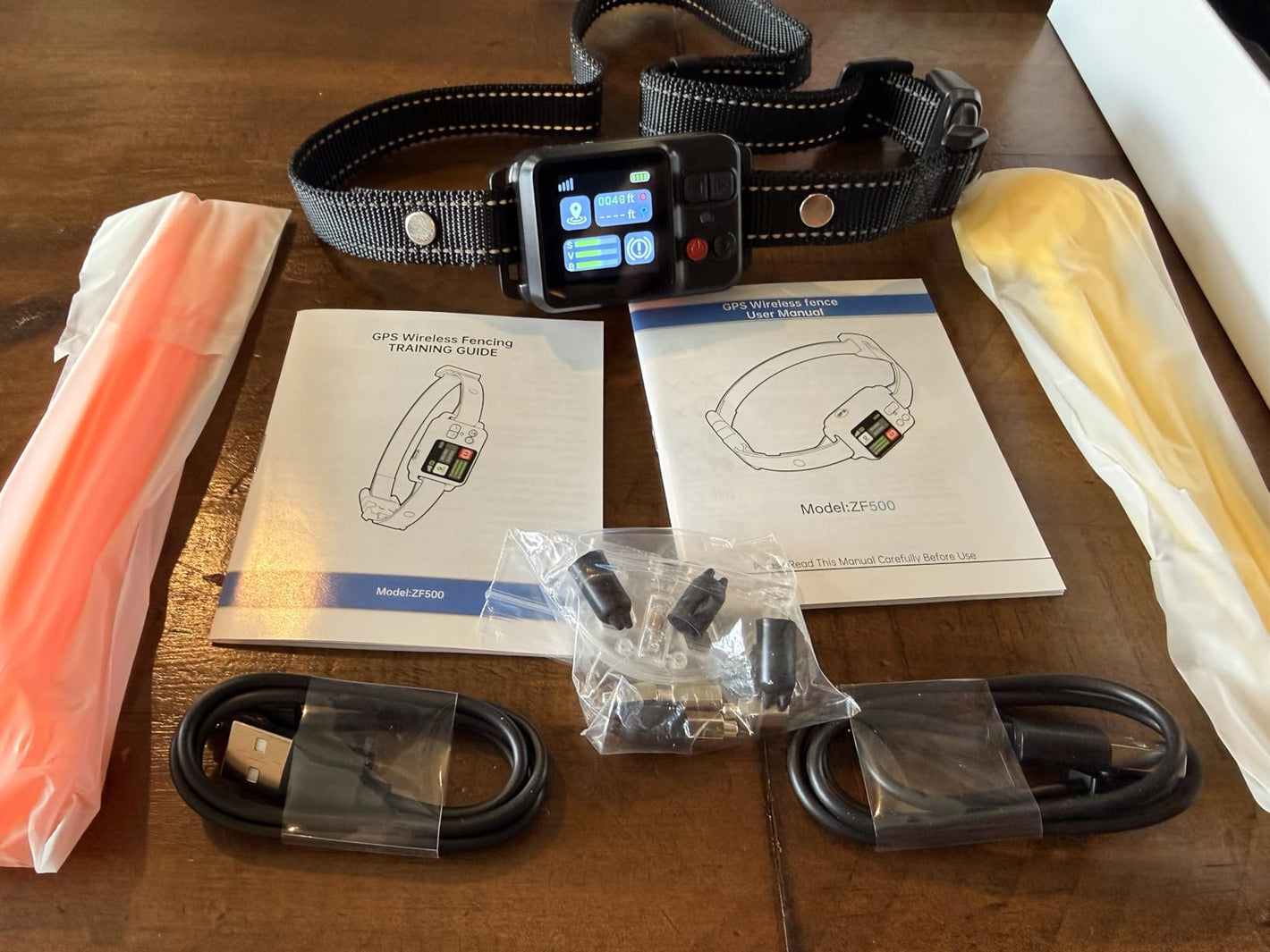 A smart pet collar with a black screen and adjustable black-and-white strap, laid out on a wooden table with cables, manuals, and accessories.